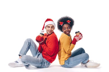 Mutlu ırklar arası çift Noel Baba şapkası ve geyik boynuzları takıyor, ellerinde kahve var, beyaz tenli.