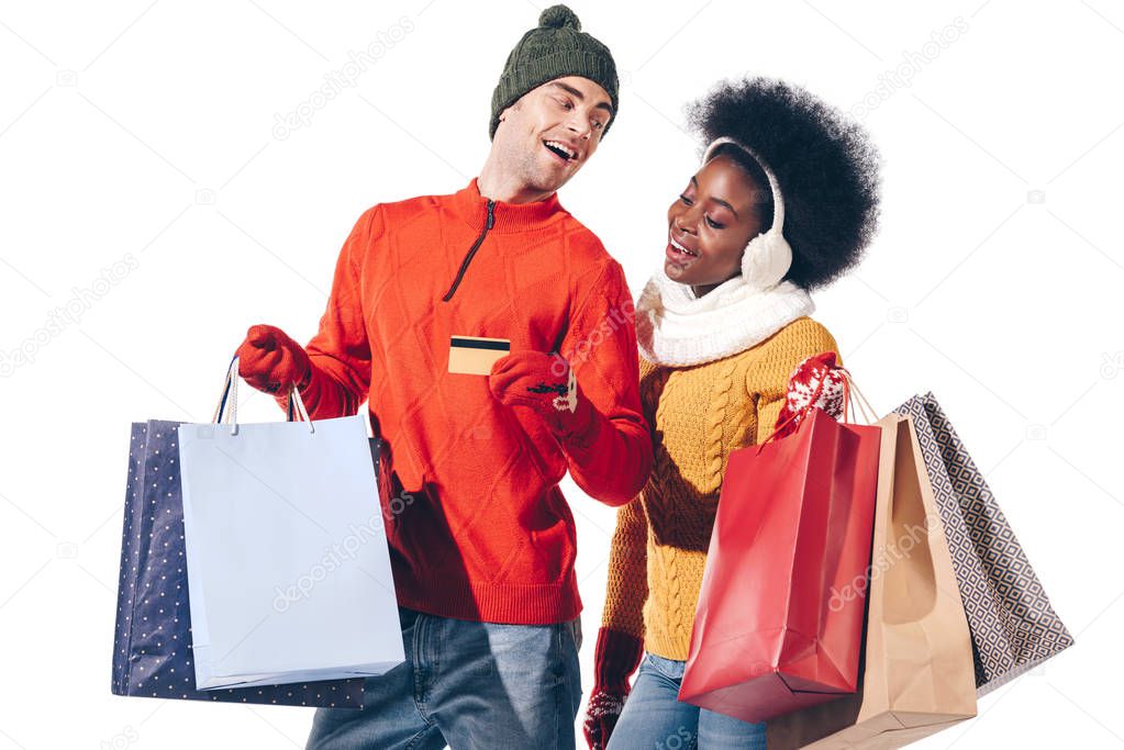 beautiful multiethnic couple in winter sweaters holding credit card and shopping bags, isolated on white