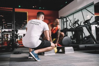 back view of personal trainer supervising african american athlete lifting weight in gym clipart