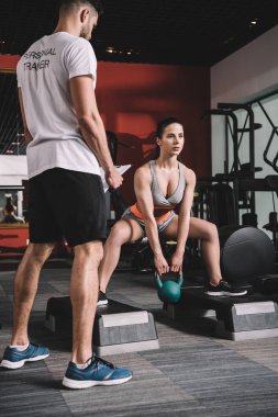 personal trainer supervising young sportswoman lifting weight in gym clipart