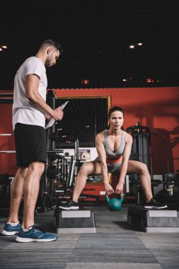 attentive trainer holding clipboard while standing near supervising young sportswoman lifting weight in gym clipart