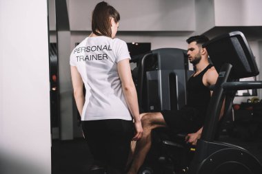 Kişisel antrenörün arka görünüşü yakışıklı sporcuyu eğitirken eliptik eğitici üzerinde çalışıyor. 