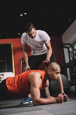 attentive trainer supervising african american sportsman doing plank exercise clipart