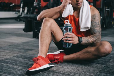 Yerde otururken elinde spor şişesi tutan yorgun Afro-Amerikan sporcunun kısmi görüntüsü 
