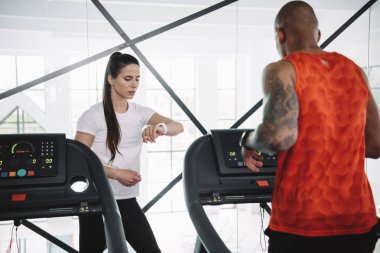 Koşu bandında koşan Afrikalı Amerikalı bir sporcunun yanında dururken fitness izleyicisine bakan dikkatli bir antrenör.