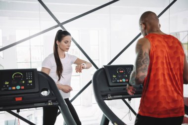 Koşu bandında koşan Afrikalı Amerikalı bir sporcunun yanında dururken fitness izleyicisine bakan dikkatli bir antrenör.