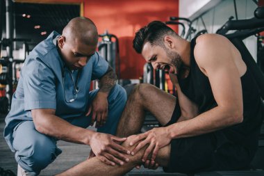 Afrikalı Amerikalı doktor bağıran sporcunun yaralı dizini inceliyor.