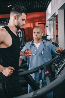 Koşu bandında koşan genç sporcuyu denetleyen ciddi bir Afrikalı Amerikalı doktor.