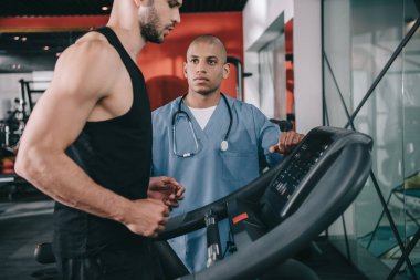 Genç Afrikalı Amerikalı doktor koşu bandında koşan sporcuyu denetliyor.
