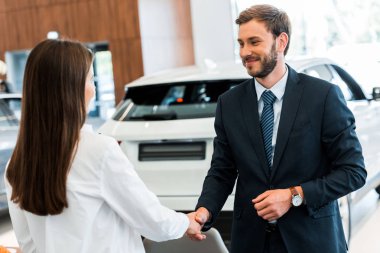 happy car dealer shaking hands with woman in car showroom 