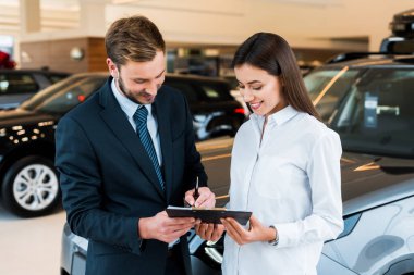 happy bearded man holding near near clipboard and woman in car showroom  clipart