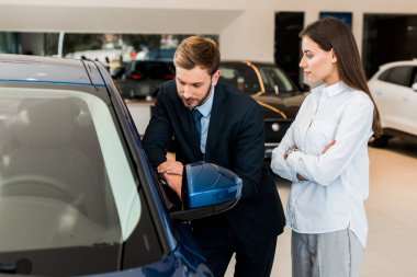 attractive woman standing with crossed arms near handsome bearded man in car showroom  clipart