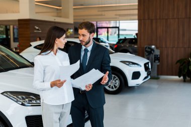 attractive woman standing with bearded man and holding papers in car showroom  clipart