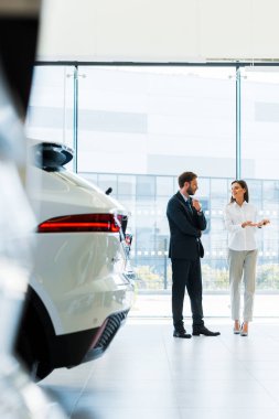 selective focus attractive car dealer gesturing near bearded man in car showroom  clipart