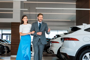 happy bearded man gesturing while walking with woman in car showroom  clipart