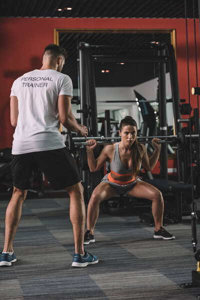 back view of personal trainer looking at young sportswoman lifting barbell