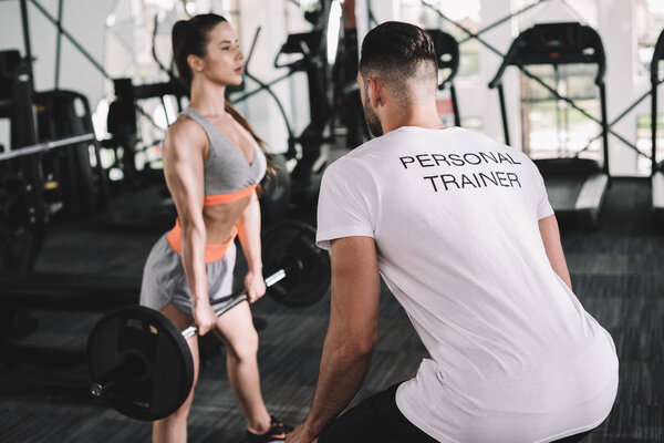back view of personal trainer looking at attractive sportswoman lifting barbell