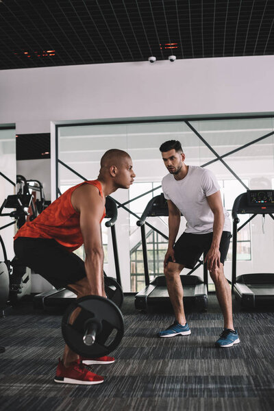 attentive trainer instructing african american sportsman lifting barbell