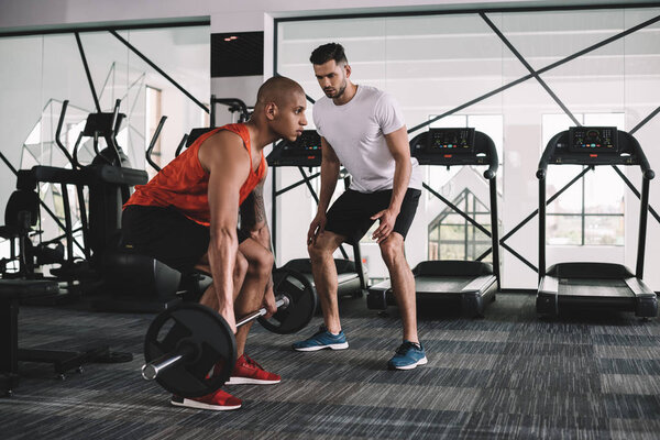 concentrated trainer looking at african american sportsman lifting barbell