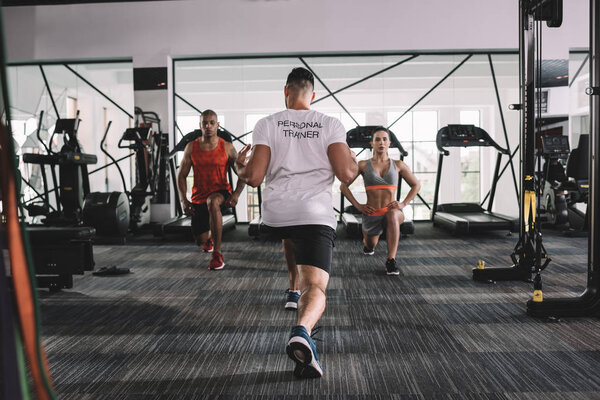 обратно вид личного тренера делать выпады упражнения вместе с мультикультурными спортсменами в тренажерном зале
