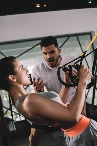 excited trainer shouting while motivating young sportswoman pulling up on suspension trainer