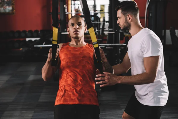 Attentive Trainer Supporting Young African American Athlete Pulling Suspension Trainer — Stock Photo, Image