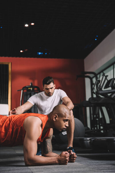 attentive trainer instructing african american sportsman doing plank exercise