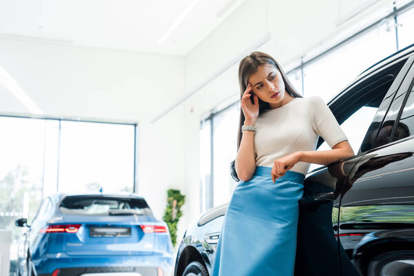 attractive and pensive woman standing near cars in car showroom 