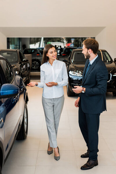 cheerful car dealer gesturing while looking at bearded man in car showroom 