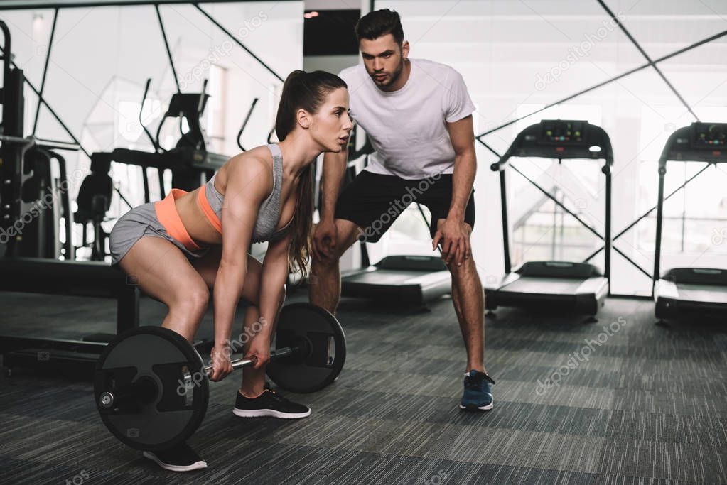 handsome trainer looking at athletic sportswoman lifting barbell