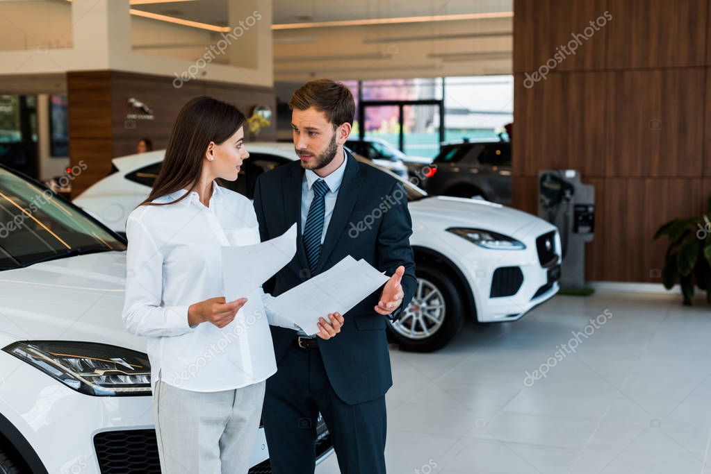 attraente donna in piedi con l'uomo barbuto e in possesso di carte in auto showroom