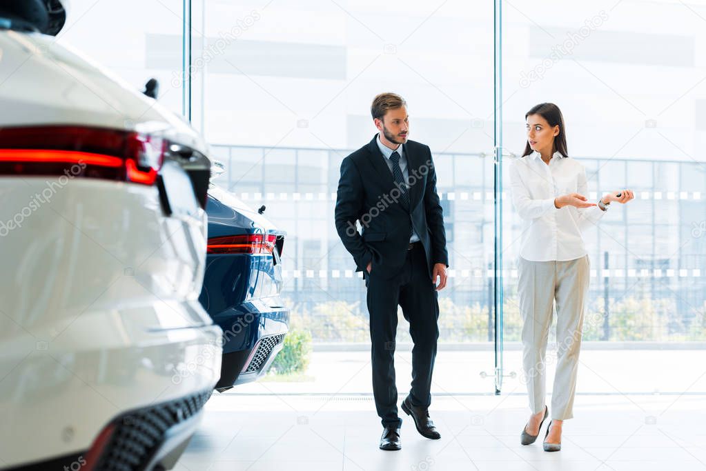 handsome man with hand in pocket walking near attractive car dealer 