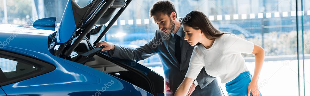 panoramic shot of woman and man looking at car trunk in car showroom 