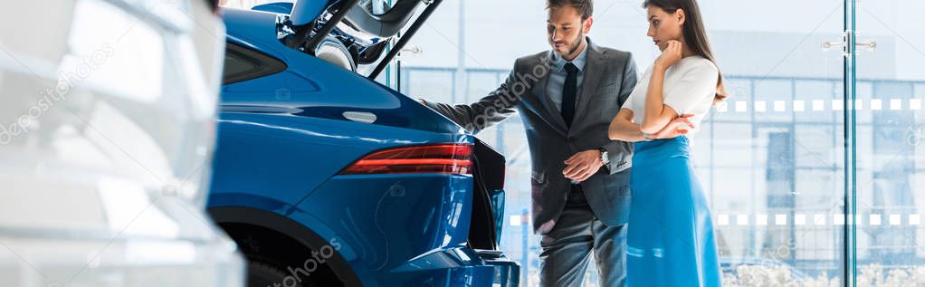 panoramic shot of attractive woman and handsome man looking at car trunk in car showroom 