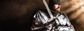 Panoramaaufnahme eines gutaussehenden Ritters in Rüstung mit Schwert auf schwarzem Hintergrund