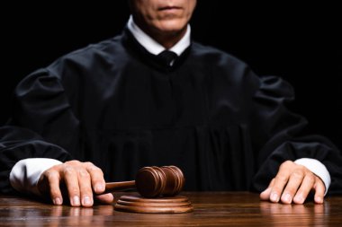 cropped view of judge in judicial robe sitting at table and holding gavel isolated on black clipart