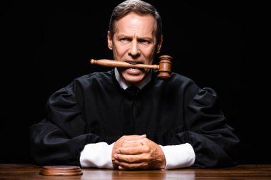 judge in judicial robe sitting at table and holding gavel in mouth isolated on black clipart