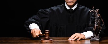 panoramic shot of judge in judicial robe sitting at table and hit with gavel isolated on black clipart