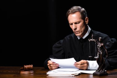 judge in judicial robe sitting at table and reading paper isolated on black clipart