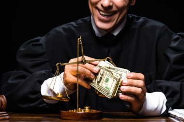 cropped view of smiling judge in judicial robe sitting at table and holding dollar banknotes isolated on black clipart