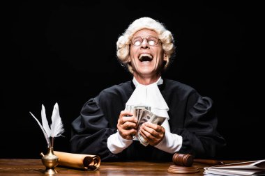 smiling judge in judicial robe and wig sitting at table and holding money isolated on black clipart