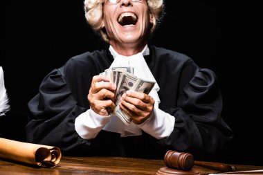 cropped view of smiling judge in judicial robe and wig sitting at table and holding money isolated on black clipart