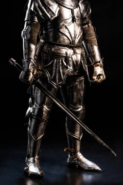 cropped view of knight in armor holding sword on black background  clipart