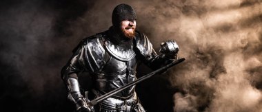 panoramic shot of handsome knight in armor holding sword and fighting on black background clipart