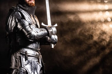 cropped view of knight in armor holding sword on black background clipart