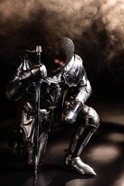 handsome knight in armor holding sword and bend knee on black background clipart