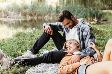 handsome man embracing girlfriend sleeping on blanket near lake in park clipart