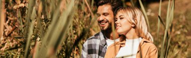 Sedge çalılığında gülümseyen mutlu genç çiftin seçici odak noktası, panoramik çekim