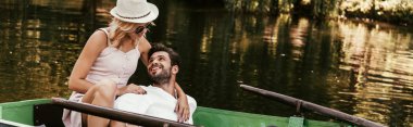 Genç bir kadının teknede otururken şapkasıyla mutlu sevgilisine sarılmasının panoramik görüntüsü.