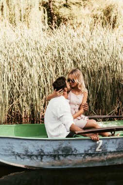 young couple embracing and kissing in boat on river near thicket of sedge clipart